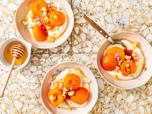 Trois bols de compote abricot onctueuse, garnie d'amandes et de fleurs violettes. Un pot de miel avec sa cuillère est à côté.