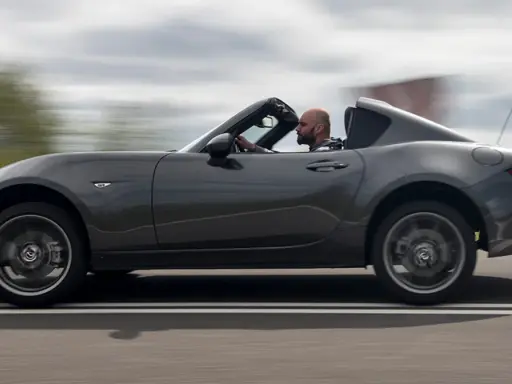 Mężczyzna w szarej kurtce prowadzi ciemnoszarą Mazdę Miata z opuszczonym dachem, jadąc po drodze.