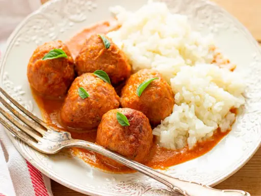 Albondigas a la jardinera con arroz blanco, decoradas con hojas verdes. Un tenedor plateado descansa sobre el plato.