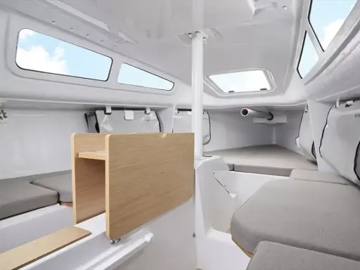 Intérieur du FIRST 24, cabine lumineuse avec banquettes grises, table en bois et hublots ouverts sur le ciel bleu.