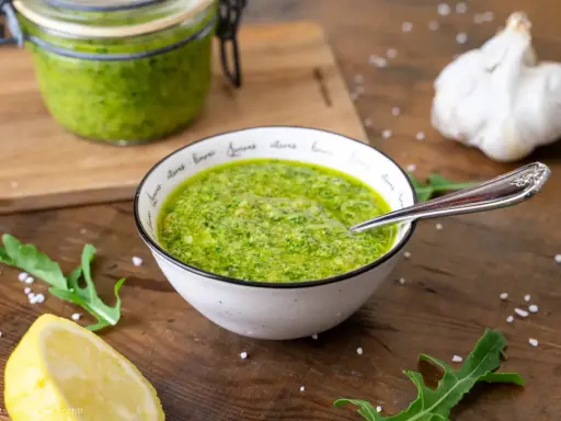 Frisches Rucola Pesto in einer Schüssel, daneben Zitrone, Knoblauch und Rucolablätter.