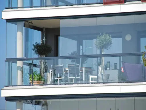 Un balcone moderno con vetrate scorrevoli, ideale per chiudere un balcone fai da te. Tavolo, sedie e un pouf viola creano un angolo relax.