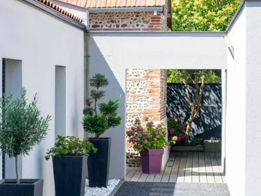 Idée aménagement jardin devant maison : olivier et conifères en pots, passage vers un patio en bois avec hortensias.