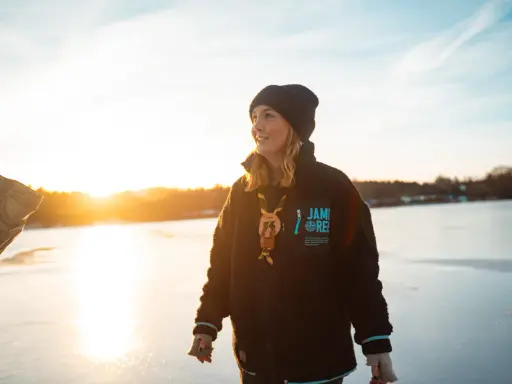 Young woman in a black beanie and fleece jacket standing on a frozen lake at sunset.