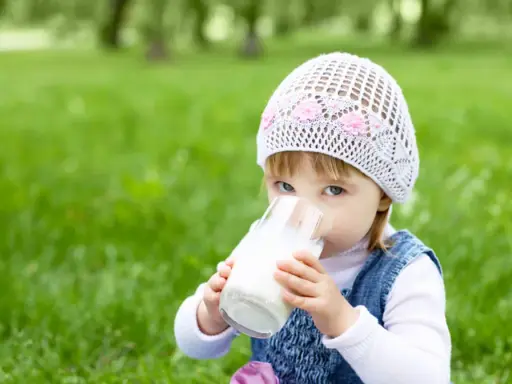 Mała dziewczynka w czapce pije mleko owsiane dla niemowlaka ze szklanki na trawie.
