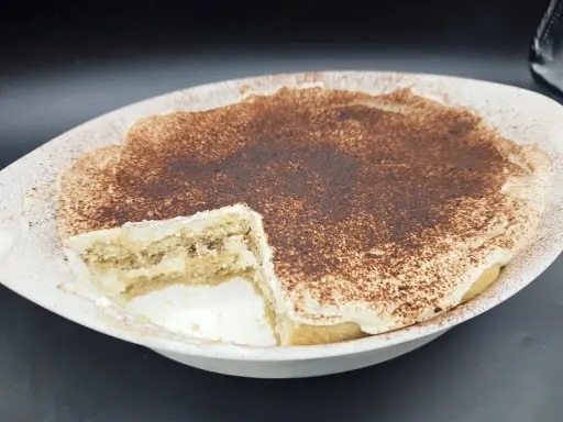 Un délicieux tiramisu sans oeuf, crémeux et saupoudré de cacao, dans un plat blanc. Une part a été retirée, révélant les couches de biscuits et de crème.