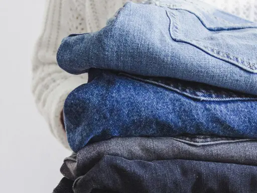 En hög med jeans i olika blå nyanser, redo för jeans återvinning. En vit stickad tröja syns i bakgrunden.