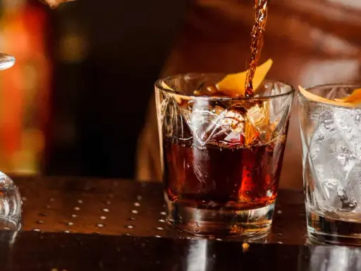 Ein Drink wird in ein Glas mit Eis und Orangenzeste gegossen. So trinkt man Whisky.
