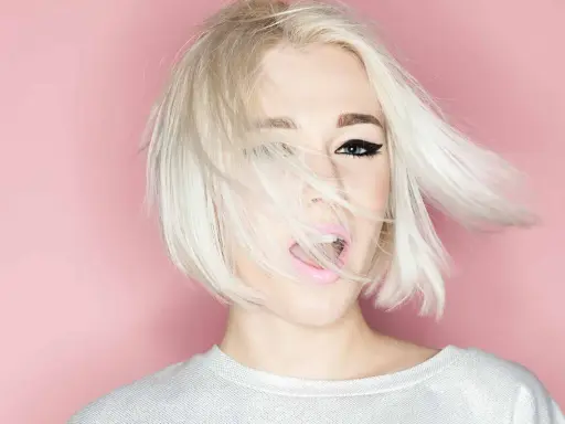 Mujer con cabello rubio platino y corte recto al hombro, con un ojo visible y labios rosados abiertos.