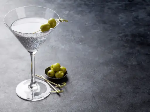 Ein Glas trockener Wermut mit Oliven garniert, daneben eine Schale mit weiteren Oliven auf dunklem Untergrund.