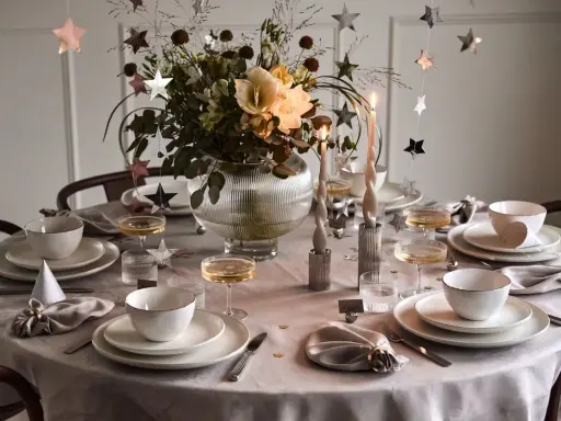 Enkel nyårsdukning med vita tallrikar, champagneglas, stjärnformade dekorationer och en blomsterbukett i en glaskaraff.