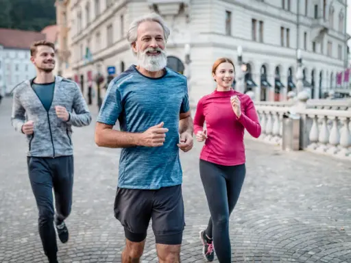 Un uomo con barba bianca e due giovani corrono sorridenti in città. Un esempio di come iniziare a correre a 50 anni sia possibile e gratificante.