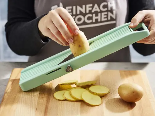 Eine Hand schneidet Kartoffeln für ein Kartoffelsandwich auf einem Hobel. Dünne Scheiben liegen auf dem Holzbrett.