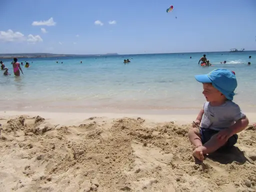 Bambino con cappellino blu gioca sulla sabbia a Cipro, mentre altri si divertono in mare.