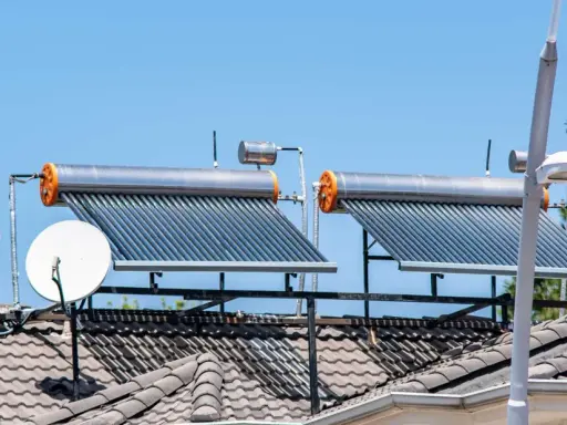 Dos calentadores de agua caliente solar en un tejado, junto a antenas parabólicas, bajo un cielo azul.
