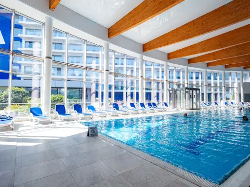 Wnętrze z basenami hotelowymi, leżakami i dużymi oknami z widokiem na budynek hotelowy.