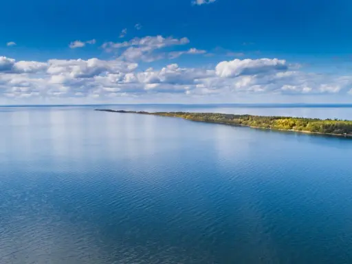 Blå himmel med moln över ett lugnt vatten. En lång, grönskande udde sträcker sig ut. Perfekt för vandring Mariestad.