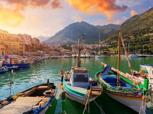 Malowniczy port w Trapani, idealne miejsce na zwiedzanie. Łodzie kołyszą się na wodzie, a w tle widać góry i kolorowe budynki.