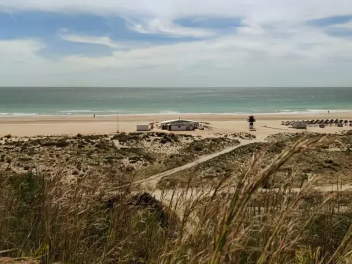 Strand von Conil de la Frontera mit Dünen, einem Strandcafé und Sonnenschirmen. Das Meer ist ruhig, der Himmel ist bewölkt.
