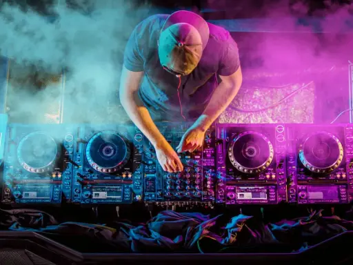 DJ con cuffie e cappellino mixa musica hause su console illuminata da luci viola e blu, circondato da fumo.