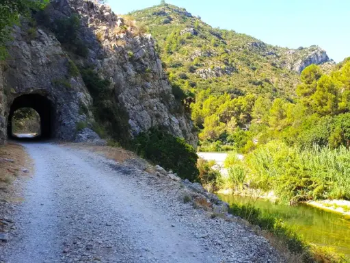 Camino de grava junto a un río y un túnel en la vía verde de Alicante. Naturaleza exuberante y rocas escarpadas.