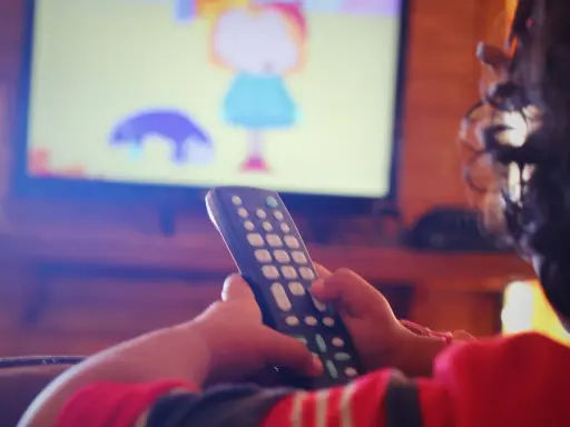 Dziecko z pilotem w ręku ogląda bajkę na telewizorze w pokoju.