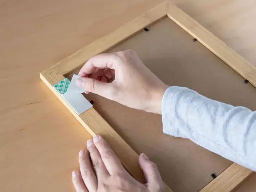 Mano retirando cinta de doble cara de un marco de madera. Aprende cómo quitar cinta de doble cara de la pared sin dañarla.