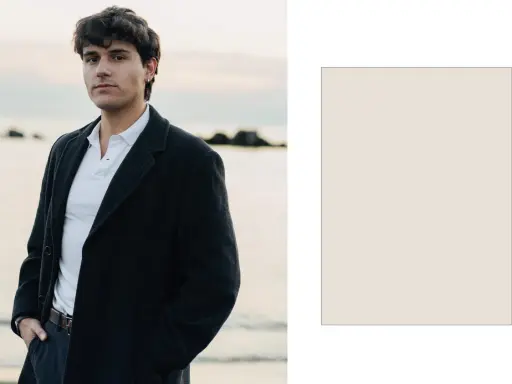 Davide Avolio, con le mani in tasca, indossa un cappotto scuro e una polo bianca, con il mare sullo sfondo.
