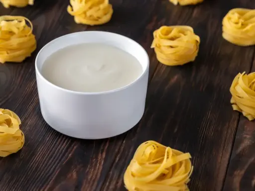 Un bol de béchamel sans beurre crémeuse, entouré de nids de pâtes fraîches sur fond de bois sombre.