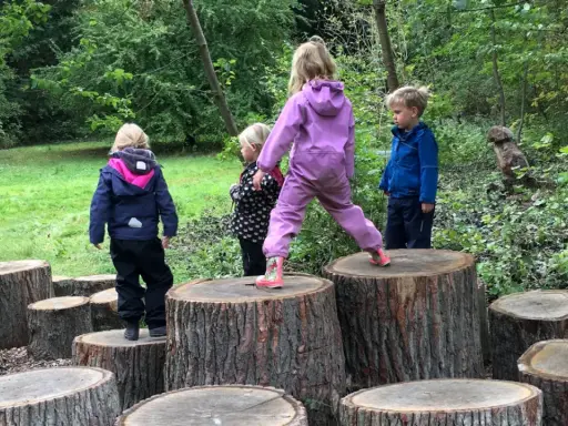 Barn i skogen leker på stubbar. En flicka i lila overall balanserar på en hög stubbe, medan andra barn tittar på eller klättrar.