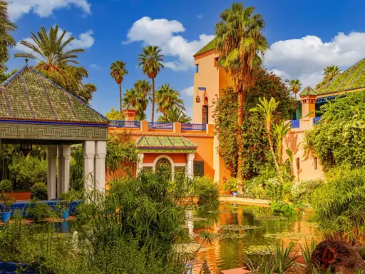 Jardin luxuriant avec un étang reflétant une maison bleue à Marrakech, palmiers et ciel bleu.