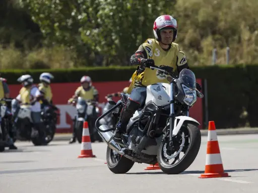 Hombre en moto blanca, con chaleco amarillo y casco, practicando maniobras en un curso carnet A. Otros motoristas y conos en el circuito.