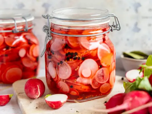Zwei Gläser mit eingelegten Radieschen, leuchtend rot und mit schwarzen Pfefferkörnern. Frische Radieschen liegen daneben.