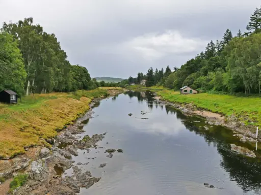 Ein ruhiger Fluss, der durch eine grüne Landschaft fließt. Kleine Häuser säumen das Ufer, vielleicht Orte, an denen man das schottische Uisge Beatha genießt.
