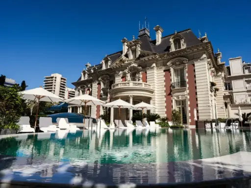 En lyxig pool framför en ståtlig byggnad som påminner om White Lotus Hotel, med solstolar och parasoller.