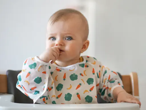 Niebieskooki maluch w śliniaczku z warzywami, kiedy dziecko rozpoznaje twarze, z paluszkiem w buzi.