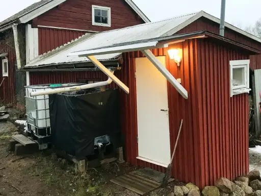 Rött utedass med vit dörr och fönster. Bredvid står en stor tank och en svart presenning. Här följer man utedass regler.