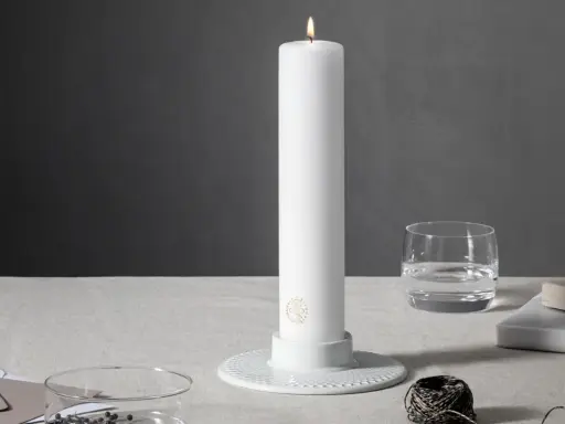 Ett vitt, högt stearinljus med guldig logotyp står i en vit ljusstake. Bredvid finns ett glas vatten, böcker och garn. Olika former av stearinljus kan skapa stämning.