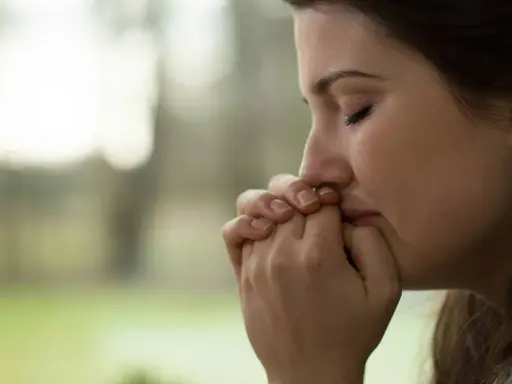 Visage pensif d'une jeune femme, mains jointes devant sa bouche, peut-être réfléchissant aux tactiques d'un manipulateur narcissique.