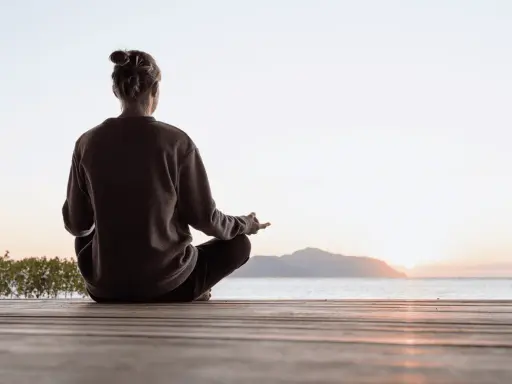 Person sitter i lotusställning på en träbrygga vid havet, med solnedgång i bakgrunden. En stund av mindful yoga.