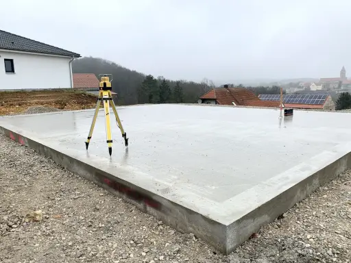 Der **Bodenplatten-Aufbau** ist fast fertig. Ein Nivelliergerät auf einem Stativ steht auf der frisch gegossenen Betonplatte. Im Hintergrund sind Häuser und eine Kirche zu sehen.