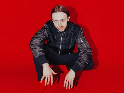 Tommy Cash w czarnej kurtce bomberce i czarnych spodniach kuca na czerwonym tle.