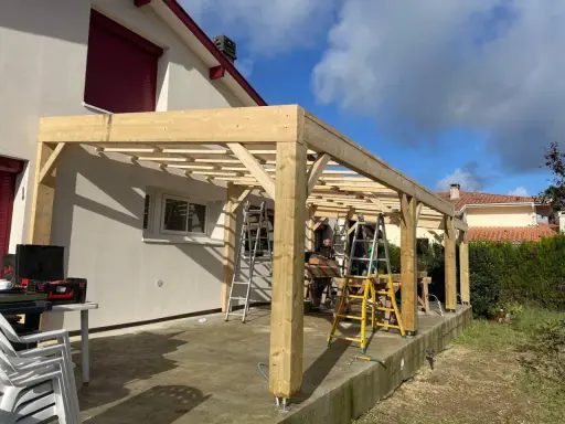 Construction d'une charpente sans ferme en bois, avec des poteaux et des poutres, sur une terrasse. Des échelles sont présentes.