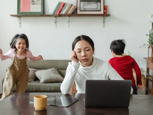 Donna esausta davanti al laptop, mentre i bambini giocano. Un quadro di **parental burnout** in casa.