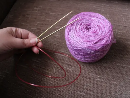 En hand håller i en rundsticka med ett nystan i lila toner bredvid. Dags att sticka resår på rundsticka!