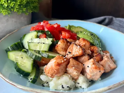 Pyszny bowl z kawałkami pieczonego lososia, świeżym ogórkiem, pomidorem i awokado na ryżu.