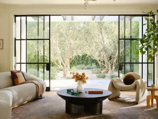 Salones modernos con sofá curvo, sillón acogedor y mesa de centro, conectados a un jardín soleado con olivos.