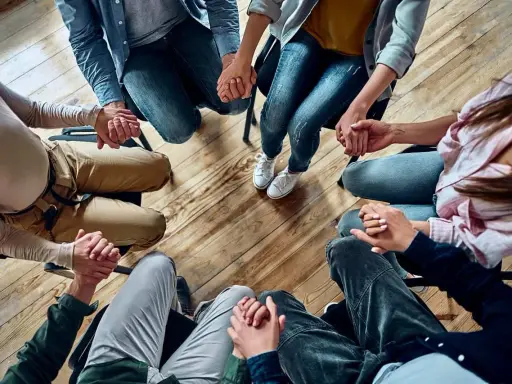 Grupa osób trzyma się za ręce w kręgu, wspierając się nawzajem. Widać tu pracę specjalisty od uzależnień.