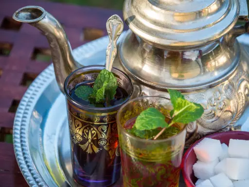 Zwei Gläser mit Minztee, garniert mit frischen Minzblättern, stehen neben einem traditionellen Teekessel und Zuckerwürfeln. Verschiedene Minze Arten verfeinern das Getränk.