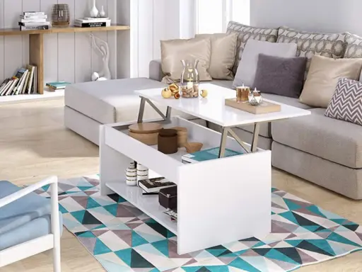 Mesa elevable pequeña blanca con compartimentos, junto a un sofá gris y una silla azul, sobre una alfombra geométrica.